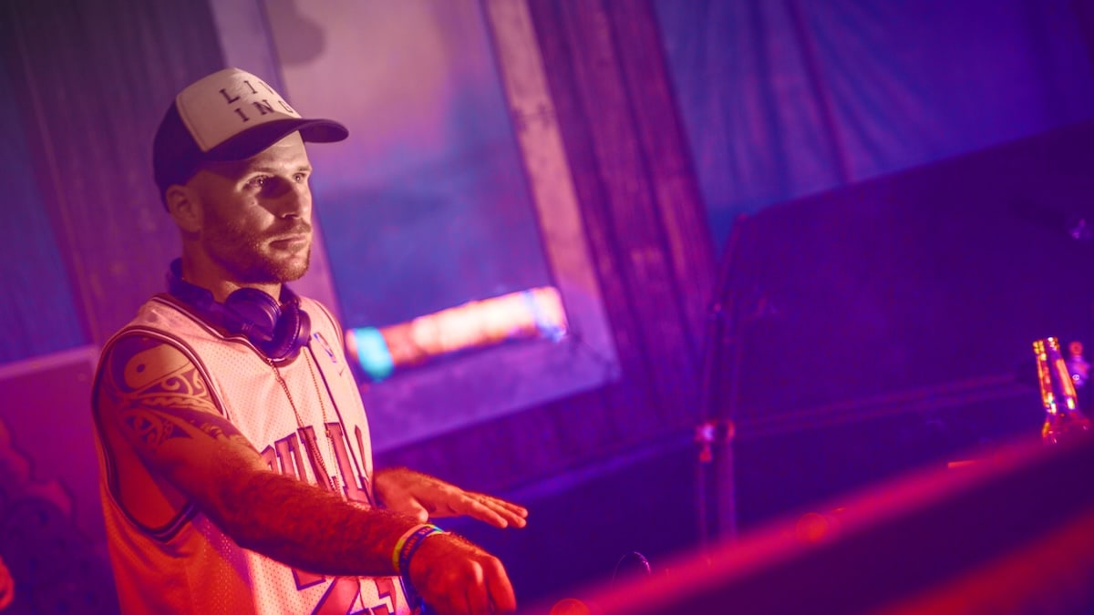 In violettem Licht steht Sascha Lebemann am DJ-Pult, trägt eine Kappe und Kopfhörer um den Hals.