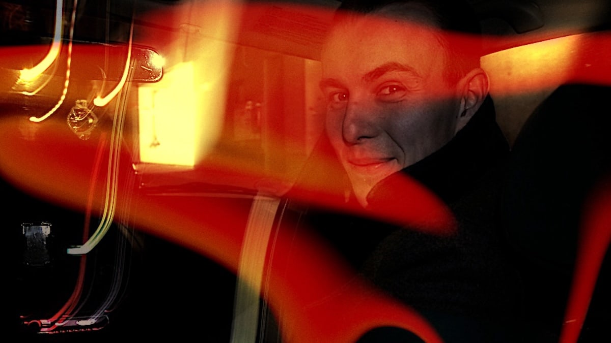 In rotem Licht blickt Thamash durch eine Autoscheibe in die Kamera; Reflexionen und dunkle Schatten prägen die Aufnahme.